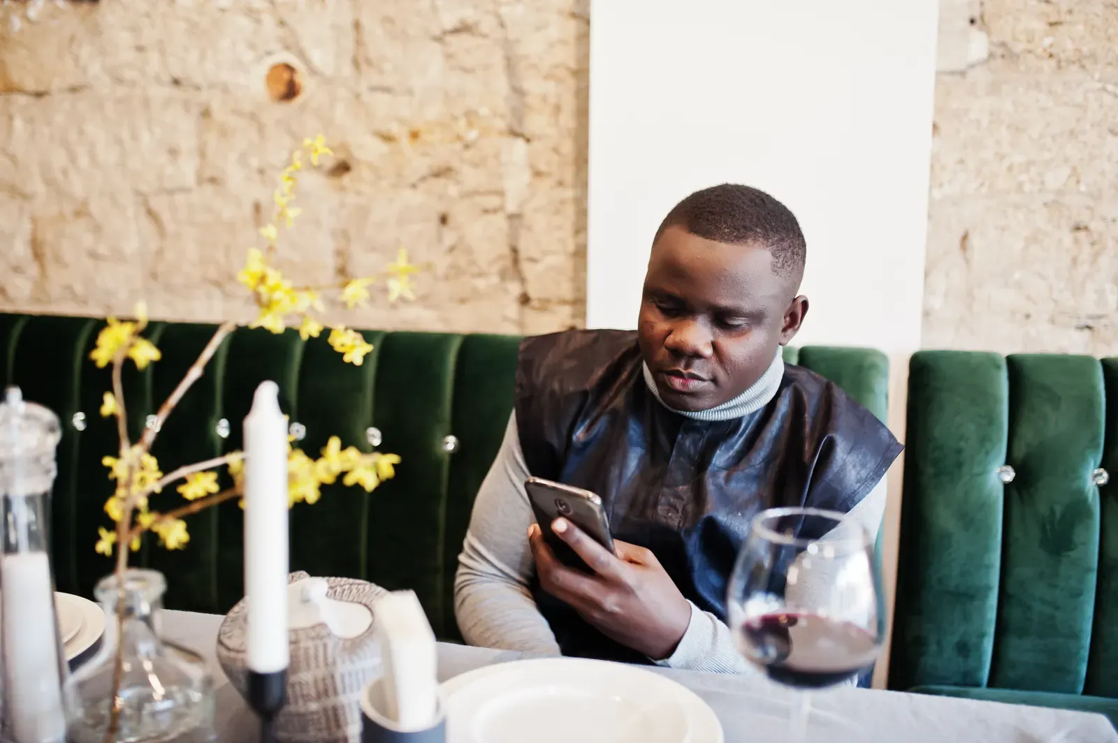 A man seated at a restaurant table looking at his phone, with a glass of red wine in front of him